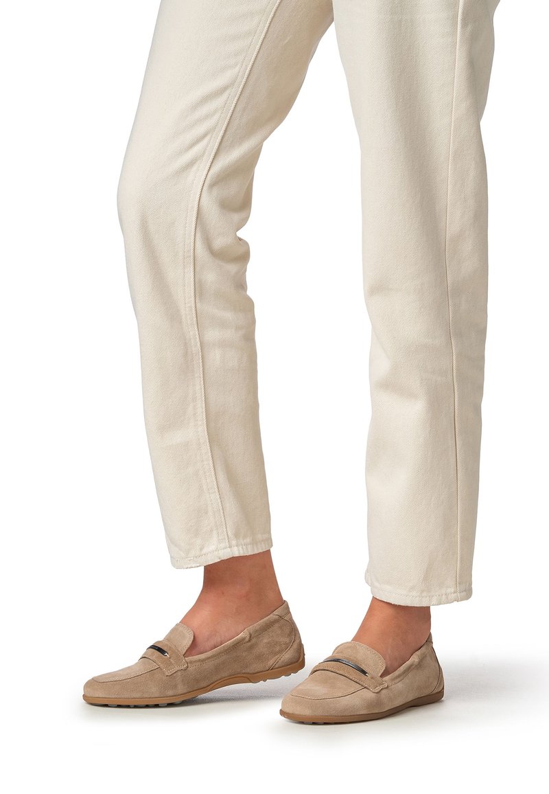 Beige Wildleder-Slipper mit flacher Sohle, mit einfachem Schnitt und einem dekorativen schwarzen Akzent oberhalb. Getragen zu hellen Hosen.