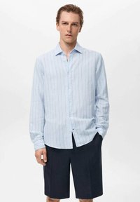 Camisa de botones de rayas azul claro con mangas largas, hecha de una tela suave, combinada con pantalones cortos oscuros. Presenta un cuello clásico y botones frontales.