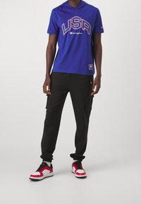 Niebieska bluza z kapturem z grafiką "USA", czarne spodnie cargo z bocznymi kieszeniami oraz czerwone i białe buty sportowe z czarnymi akcentami.