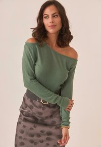 Top de punto acanalado verde sin hombros combinado con una falda gris estampada que presenta un diseño floral negro y un cinturón negro ancho con hebilla circular.