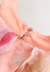 Primer plano de un escote de tela color melocotón con dos botones redondos brillantes y un tejido floral suave y translúcido debajo.
