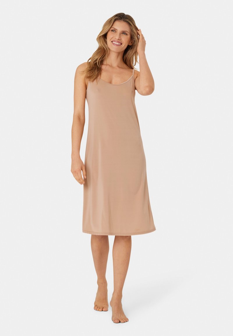 Femme souriante, debout pieds nus, portant une robe beige sans manches jusqu'aux genoux, sur un fond clair uni.