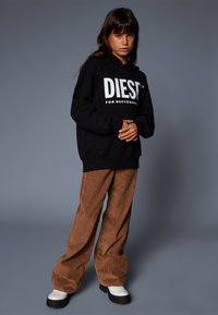 Black hooded sweatshirt with large white logo, paired with brown corduroy wide-leg pants and white platform shoes. Textured fabric and relaxed fit.