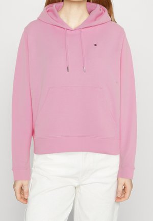 Personne portant un sweat à capuche rose avec une poche avant et un petit logo sur la poitrine, associé à un pantalon blanc.