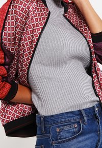 Red patterned bomber jacket with black trim, paired with a silver ribbed turtleneck. Blue denim jeans complete the outfit.