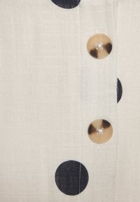 Hellfarbener Stoff mit großen marineblauen Punkten und zwei transparenten Knöpfen entlang der Vorderseite. Strukturiertes Material mit leinenartigem Aussehen.