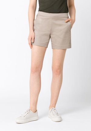 Kvinde iført beige shorts med sidelommer, en mørkegrøn top og hvide snøresneakers, stående med den ene hånd i lommen.
