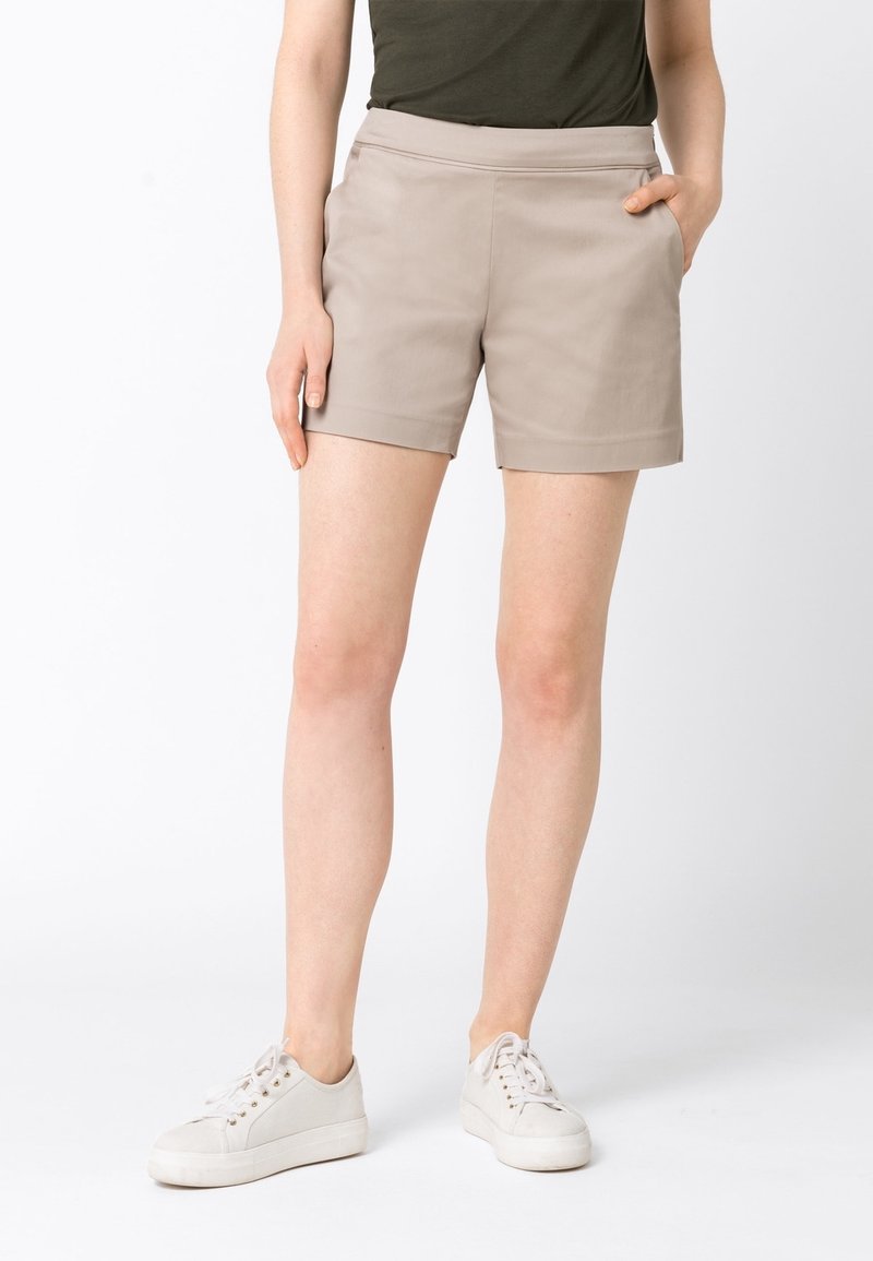 Femme portant un short beige avec des poches latérales, un haut vert foncé et des baskets blanches à lacets, debout avec une main dans la poche.