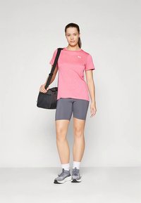 Kobieta w różowej sportowej koszulce i szarych szortach, niosąca czarną torbę na siłownię na ramieniu, stojąca na jednolitym tle.