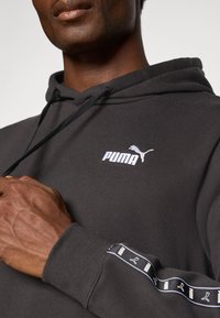 Zwarte hoodie met een trekkoord, voorzien van een wit Puma-logo op de borst. De mouw heeft een patroonaccent met een herhalend logo.