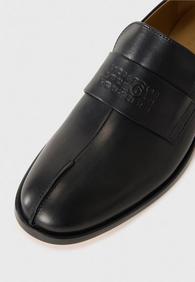 Chaussure en cuir noir au design élégant, dotée d'une sangle proéminente avec des chiffres en relief et d'une surface lisse, d'un bout arrondi et d'une semelle plate.