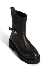 Black leather ankle boot with a chunky, ridged sole, side zip closure, and a decorative strap with metallic hardware. Smooth texture.