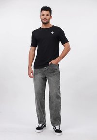 Must lühikeste varrukatega t-särk kolju logoga, millega on kombineeritud tuhmunud hallid teksad ja mustad tossud, seisab heledal taustal.