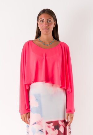 Donna con lunghi capelli scuri indossa una maglia larga rosa acceso, una gonna floreale pastello e una collana blu a forma di stella, in piedi davanti a uno sfondo bianco.