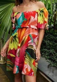 Robe décolletée avec un motif floral vibrant dans des rouges, des verts et des jaunes, ornée d'un volant à la taille et en tissu léger.