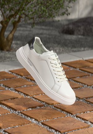 Baskets en cuir blanc avec languette de talon noire flottant au-dessus de carreaux carrés en bois avec de petites pierres grises entre eux.