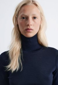 Navyblauer Rollkragenpullover aus weichem, geripptem Material mit taillierter Passform. Das Model hat glattes, hellblondes Haar, das das Gesicht umrahmt.