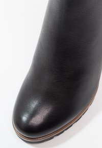 Gros plan sur l'avant d'une botte en cuir noir avec une finition lisse et une bordure marron le long du bord de la semelle sur fond blanc.