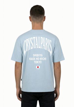 Uomo che indossa una maglietta azzurro chiaro con la scritta "CRYSTAL PARIS", "SHIBUYA KAGE NO MICHI TOKYO" e una piccola bandiera giapponese sulla schiena.
