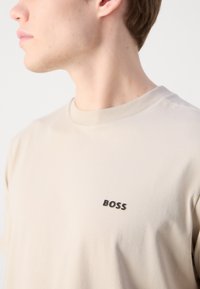Beige bomulds t-shirt med rund hals, med et sort "BOSS" logo på venstre side. Glat tekstur og afslappet pasform.