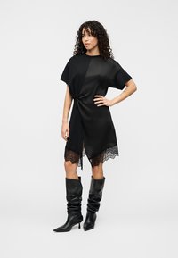 Μαύρο φόρεμα με κοντά μανίκια, λεπτομέρεια με κόμπο μπροστά, δαντελωτό τελείωμα και ψηλή σχισμή. Συνδυασμένο με μαύρες μπότες μέχρι το γόνατο, διπλωμένες.