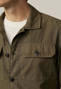 Camisa verde oliva de botones, hecha de tela duradera, con cuello amplio, dos bolsillos en el pecho y botones negros. Textura suave.