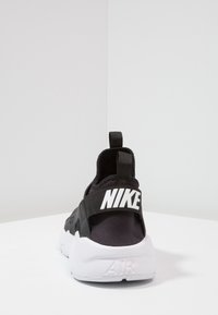 Nike shoe featuring a black fabric upper, white rubber sole, and prominent black strap with white logo. Back view shows a cushioned collar.