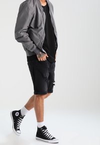 Persona con chaqueta gris, camiseta negra, pantalones cortos negros rasgados, calcetines blancos y zapatillas altas negras en medio de un paso sobre un fondo blanco.