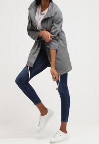 Šedá lehká bunda s kapucí, s knoflíkovými detaily a stahovací šňůrkou. Nositá přes bílý tričko a spárovaná s tmavě modrými úzkými džínami a bílými teniskami.