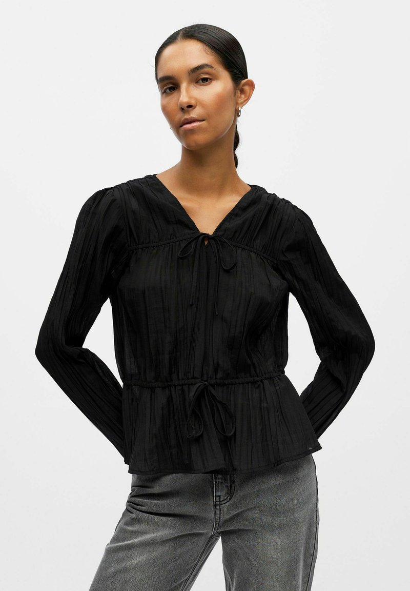 Blusa nera a maniche lunghe con trama plissettata, scollatura a V, vita con coulisse e maniche a sbuffo, abbinata a jeans grigi.