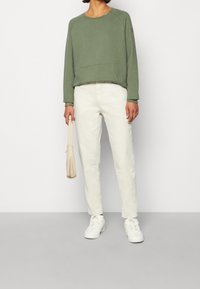Sudadera verde con un bolsillo frontal, combinada con vaqueros rectos de color blanco roto y zapatillas blancas. Los accesorios incluyen un bolso de color beige claro.