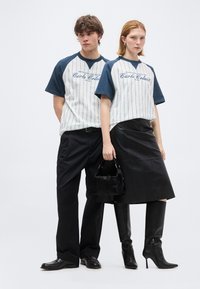 Νέος άνδρας και γυναίκα φοράνε τυπωτά T-shirts σε λευκό και μπλε ναυτικό χρώμα που ταιριάζουν· η γυναίκα κρατά μαύρη τσάντα χειρός, φορά μαύρη φούστα και μπότες μέχρι το γόνατο.