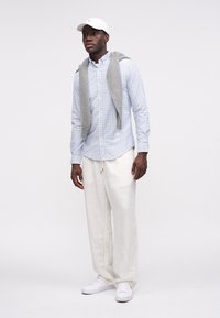 Μοντέλο που φοράει ανοιχτό μπλε πουκάμισο με καρό σχέδιο και κουμπιά, γκρι πουλόβερ περασμένο στους ώμους, μπεζ παντελόνι από λινό και λευκά αθλητικά παπούτσια.