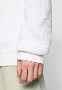 Biały prążkowany materiał swetra o teksturowanej powierzchni. Mankiet jest szeroki i posiada dziergany wzór. Ręka spoczywa na jasnym materiale.