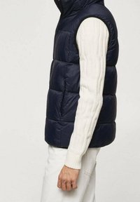 Tummansininen toppaliivi, jossa on korkea kaulus, tikkaukset ja vetoketjukiinnitys, puettuna vaalean kermanvärisen villapaidan päälle, jossa on palmikkoneule.