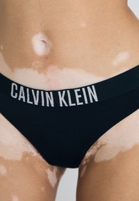 Crna Calvin Klein donji veš s širokom elastičnom trakom koja sadrži bijelo slovo. Koža s različitim tonovima vidljiva oko struka.