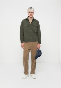 Green button-up shirt with two chest pockets, tan corduroy pants, and white sneakers; holding a blue cap with a patterned texture.