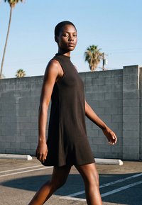 Nő ujjatlan fekete ruhában, szabadtéren sétál egy szürke betonfal mellett, háttérben pálmafák és tiszta égbolt.