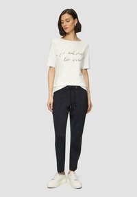 Witte korte mouwen t-shirt met gouden tekst, gecombineerd met zwarte tapered pants met een trekkoord. Gecombineerd met witte sneakers op een grijze achtergrond.