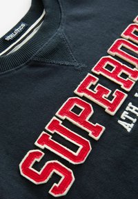 Námořnická mikina s velkým vyšitým textem "SUPERDRY" v červené a bílé barvě, kulatým výstřihem a žebrovanými manžetami. Měkký materiál s ležérním designem.