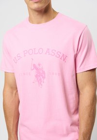 Rožinė medvilninė marškinė su trumpomis rankovėmis. Puošia U.S. Polo Assn. tekstas ir polo žaidėjo grafika šviesesnėje rožinėje spalvoje.