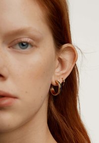 Pendientes tipo aro en oro y negro con un detallado texturizado y un pequeño pendiente en la oreja de un modelo, complementados por cabello castaño rojizo y piel suave.