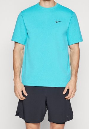 Mężczyzna w turkusowej koszulce Nike i czarnych spodenkach Nike, stojący z ramionami swobodnie opuszczonymi wzdłuż ciała.