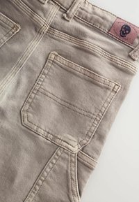 Jean en denim gris avec une poche arrière standard à double couture. Comprend un patch de marque avec un design de crâne. Texture lisse.
