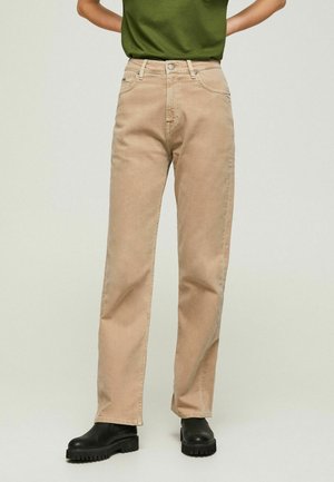 Osoba w beżowych spodniach o prostych nogawkach, zielonym topie i czarnych butach na grubej podeszwie, stojąca z rękami na biodrach.