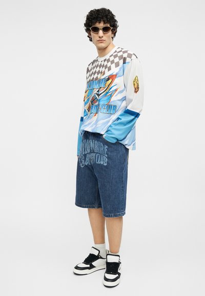 Άνδρας με καρό και γραφικό πουκάμισο μακρυμάνικο, μπλε τζιν σορτς με κείμενο, μαύρα και λευκά αθλητικά παπούτσια και γυαλιά ηλίου, στέκεται μπροστά σε λευκό φόντο.