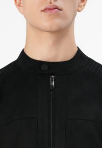 Primer plano del cuello y la parte inferior de la cara de una persona que lleva una chaqueta negra con cremallera y un cuello con botón a presión.