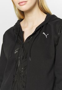 Hoodie zippé noir avec un design texturé, comportant un logo argenté, une capuche à cordon de serrage et un mélange d'accents lisses et brillants.