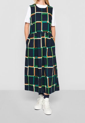 Kvinde iført navyblå ærmeløs kjole med grønt, orange og hvidt ternet mønster over en hvid T-shirt og hvide høje sneakers.