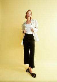 Lichtbeige blazer over een witte geribde tanktop, gecombineerd met zwarte wijde broek en zwarte slide-sandalen, tegen een gele achtergrond.
